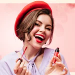 A woman wearing a red beret and lavender blouse applies red lipstick, smiling in front of a pink floral background.