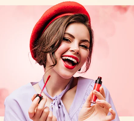A woman wearing a red beret and lavender blouse applies red lipstick, smiling in front of a pink floral background.
