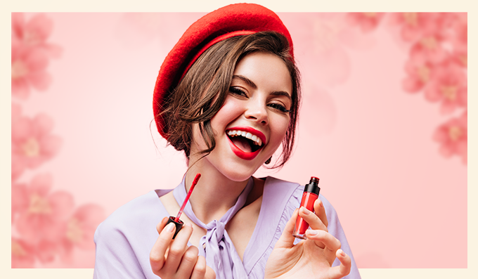 A woman wearing a red beret and lavender blouse applies red lipstick, smiling in front of a pink floral background.