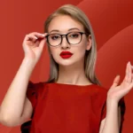 A woman with blonde hair wears glasses and bold red lipstick, set against a rich red background. She gazes confidently, touching her frames, exuding elegance.