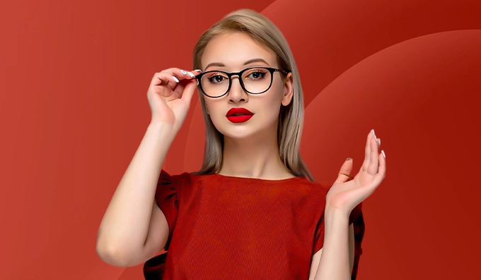 A woman with blonde hair wears glasses and bold red lipstick, set against a rich red background. She gazes confidently, touching her frames, exuding elegance.